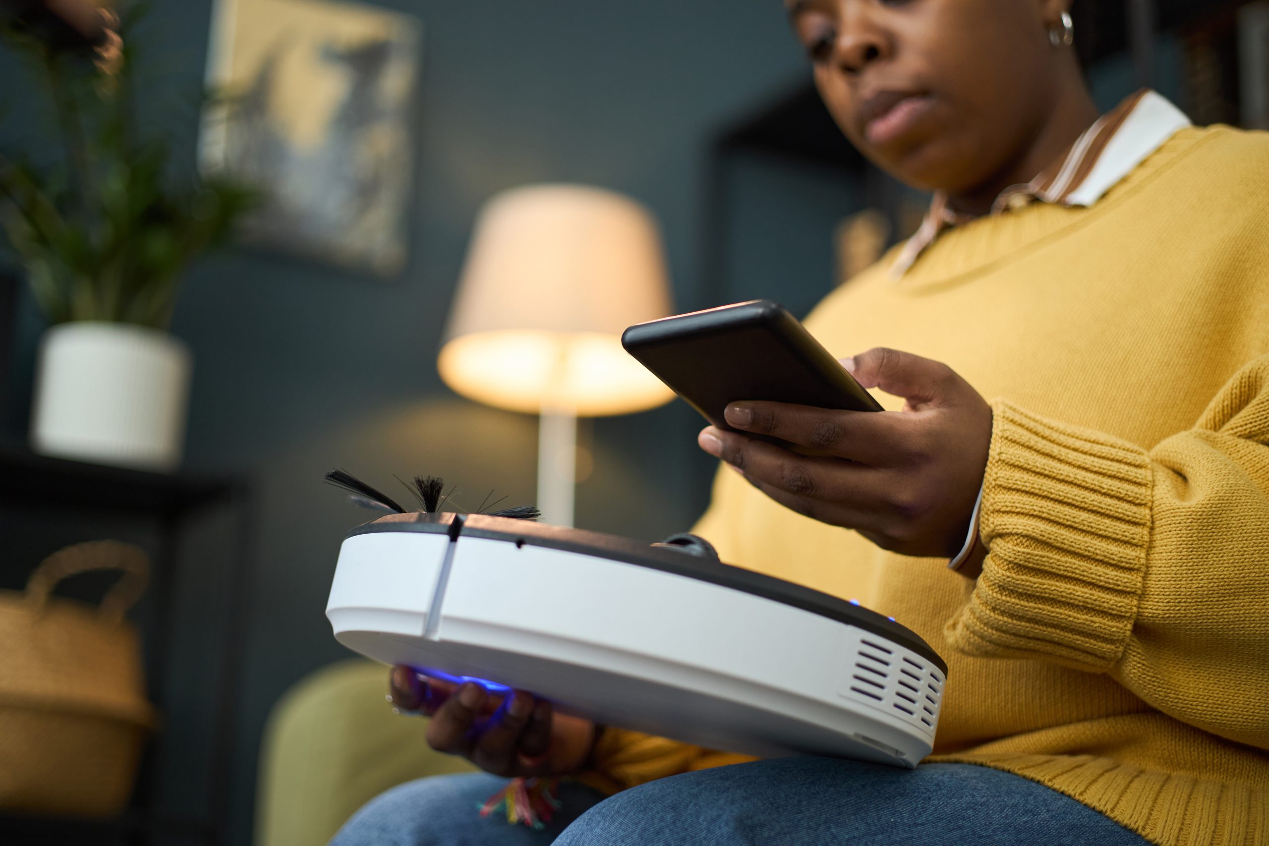 Woman sitting in living room, using smartphone to operate smart vacuum cleaner